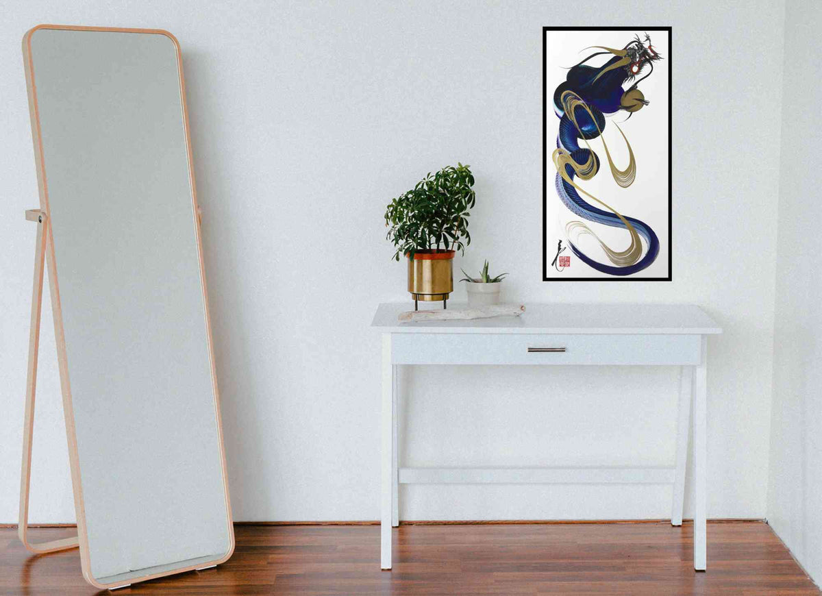 A framed abstract art print featuring a stylized blue, gold, and white dragon hangs on a white wall above a white console table with a potted plant. To the left of the table, a tall floor mirror with a light wood frame leans against the wall on a dark wood floor.