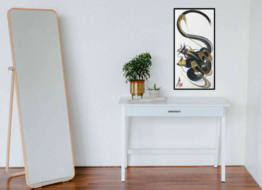 A framed vertical wall art print featuring an abstract black, gray, and gold snake-like design, displayed on a white console table in a minimalist room next to a tall floor mirror.